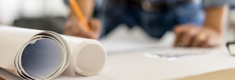 Person zeichnet auf einem Tisch mit gerollten Bauplänen im Vordergrund