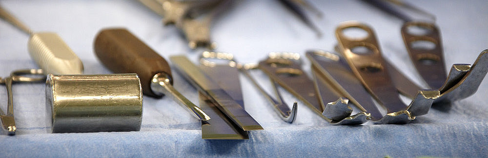Chirurgische Instrumente wie Skalpelle, Haken und Scheren auf einem blauen Tuch, im Hintergrund eine Hand mit Handschuh